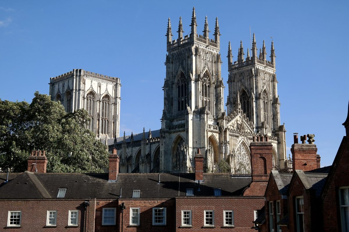 The towers of York Minster rising above red-brick buildings, seen from the city walls under a blue sky
